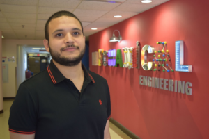 Ali Rabeh standing in front of a wall with the word ‘ENGINEERING’ in large, bold letters, wearing a dark polo shirt with red accents on the collar and sleeves.